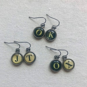 May include: Four pairs of earrings featuring typewriter keys with the letters J, T, O, K, and X. The keys are black with yellow letters and are attached to silver hooks.