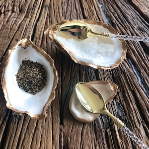 May include: Two white oyster shell dishes with gold accents, each with a gold spoon. One dish contains black peppercorns and the other is empty.