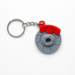 Könnte beinhalten: Ein Schlüsselanhänger bestehend aus einer grauen, kreisförmigen, perforierten Bremsscheibe und einem leuchtend roten Bremssattel. Die Teile sind durch einen silberfarbenen Metallring und eine kurze Kette verbunden, ein detailliertes Zubehör.