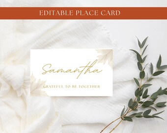 Puede incluir: Una tarjeta de sitio rectangular blanca con el nombre Samantha en letra cursiva dorada, el texto GRATEFUL TO BE TOGETHER en letras mayúsculas doradas e ilustraciones de hojas de arce beige pálido en las esquinas de la tarjeta.