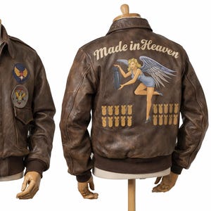 Könnte beinhalten: Braune Leder-Bomberjacke mit klassischem Kragen, Vordertaschen und Aufnähern. Auf der Rückseite befindet sich ein gemaltes Design mit den Worten "Made in Heaven" und einer Pin-up-Girl-Illustration. Die Jacke hat einen gerippten Saum und Bündchen.