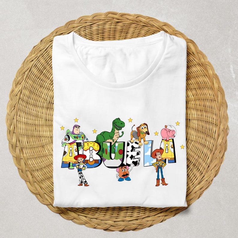 May include: White t-shirt featuring the word "BUELA" in colorful letters with cartoon characters. The design includes characters like Buzz Lightyear, Rex, and Woody, with yellow stars scattered around the letters. The shirt is folded on a woven surface.