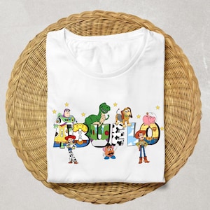 May include: White t-shirt featuring the word "ABUELO" in colorful, cartoon-style letters with Toy Story characters. The design includes Buzz Lightyear, Rex, Slinky Dog, Hamm, and Woody. The shirt is folded on a woven wicker surface.