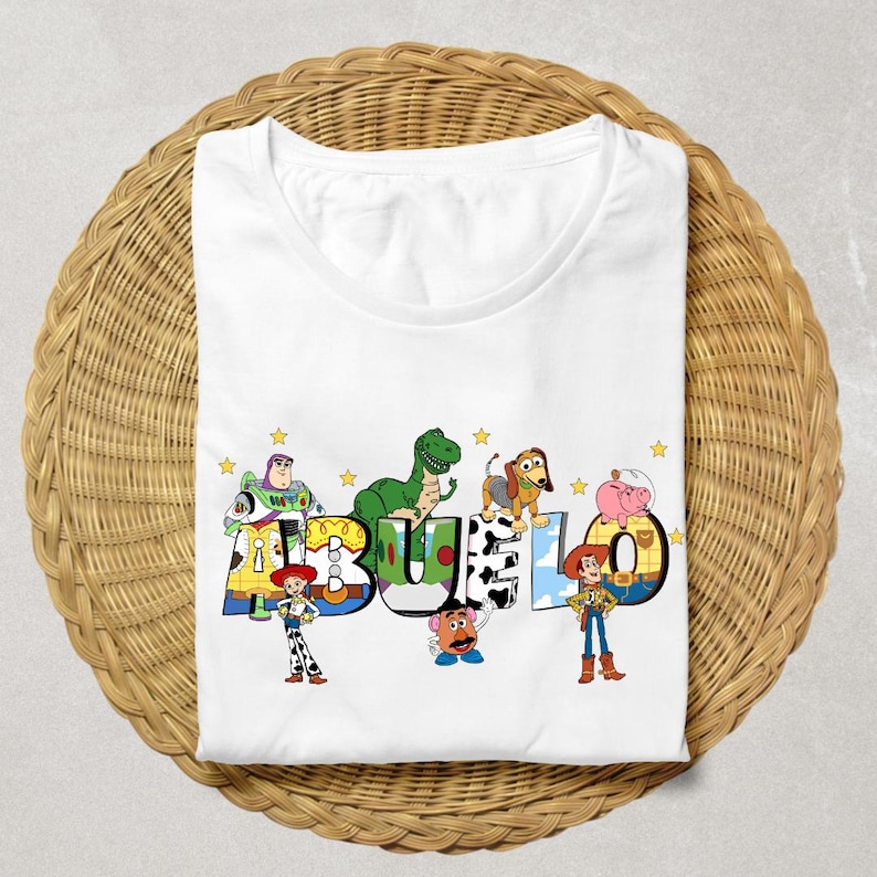 May include: White t-shirt featuring the word "ABUELO" in colorful, cartoon-style letters with Toy Story characters. The design includes Buzz Lightyear, Rex, Slinky Dog, Hamm, and Woody. The shirt is folded on a woven wicker surface.