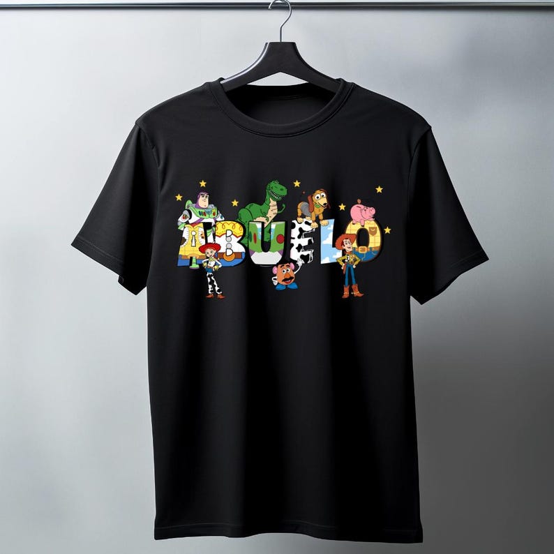 May include: Black crew neck t-shirt featuring the word "BUEL" in colorful, cartoon-style lettering. The letters are designed with characters from a popular animated movie, with yellow stars scattered around the letters.