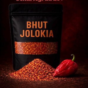 Könnte beinhalten: Ein schwarzer Beutel mit Bhut Jolokia Chili-Flocken mit dem Text "BHUT JOLOKIA" in Orange. Ein Haufen roter Chili-Flocken und eine ganze rote Chilischote befinden sich vor dem Beutel. Der Text "Schärfegrad 10+" steht oben.