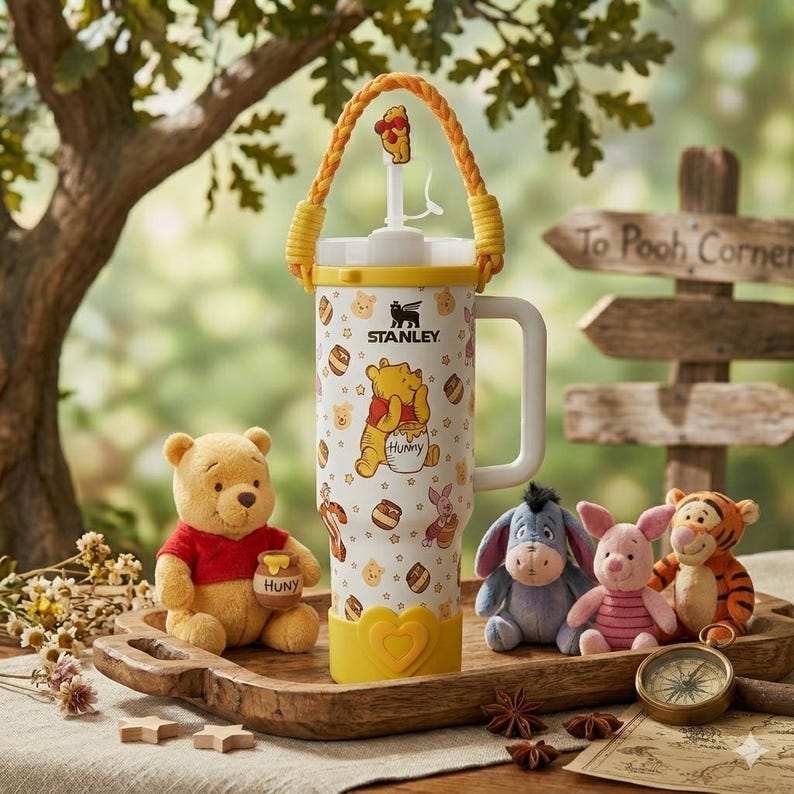 Puede incluir: Un vaso Stanley blanco con un estampado de Winnie the Pooh con tarros de miel y personajes, asa amarilla, un adorno de paja trenzado amarillo con una figura de Pooh y una base amarilla en forma de coraz&oacute;n, rodeado de peluches.