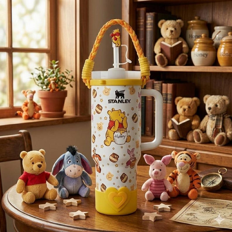 Puede incluir: Un vaso Stanley blanco con un patr&oacute;n de Winnie the Pooh, tarros de miel y personajes. Tiene un asa trenzada amarilla, una base amarilla en forma de coraz&oacute;n, una tapa blanca y un adorno para la pajita con forma de Pooh. El logo Stanley es negro.