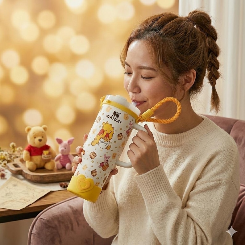 Puede incluir: Un vaso Stanley blanco con un patr&oacute;n repetitivo de Winnie the Pooh y Piglet, tarros de miel y peque&ntilde;as flores. El vaso incluye una tapa amarilla, una correa de transporte trenzada amarilla, un asa blanca y una base amarilla en forma de coraz&oacute;n.