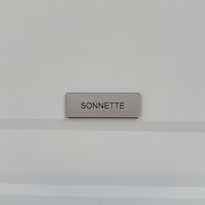 May include: A small, rectangular silver nameplate with the word "SONNETTE" in black lettering. The nameplate is affixed to a white surface. The text is centered.