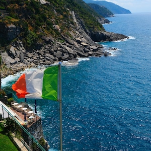 Puede incluir: Bandera italiana ondeando al viento contra un telón de fondo de costa rocosa y mar azul profundo. Un restaurante con mesas y sombrillas es visible en una terraza. La exuberante vegetación verde cubre la ladera.