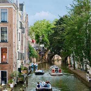 Puede incluir: Una vista pintoresca de un canal en una ciudad europea, con barcos que transportan pasajeros. Los edificios bordean el curso de agua, y un puente se arquea sobre el agua. Los árboles verdes y el cielo azul completan la escena.