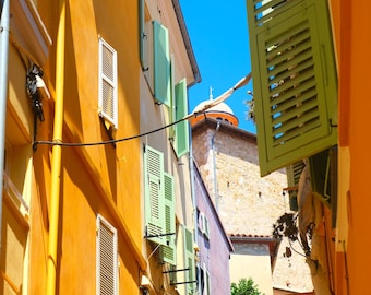 May include: A vibrant street scene with buildings painted in shades of orange, yellow, and purple. Green shutters adorn the windows, and a clear blue sky fills the space between the buildings. A church dome is visible in the distance.