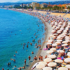 May include: A vibrant beach scene with clear turquoise water and a pebbled shoreline. Numerous people are swimming and sunbathing under white umbrellas. Buildings and palm trees line the coast under a bright blue sky.