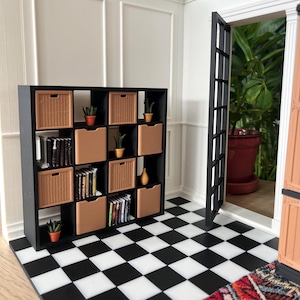 Könnte beinhalten: Ein quadratisches Miniatur-Regal in Schwarz mit sechzehn Fächern, ausgestattet mit beigen geflochtenen Körben, einfarbigen beigen Aufbewahrungsboxen, kleinen grünen Topfpflanzen und Reihen winziger Bücher auf einem schwarz-weiß karierten Boden.