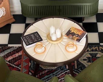 Könnte beinhalten: Ein runder weißer Couchtisch mit Strahlenmuster, darauf zwei Miniaturbücher mit den Titeln Ninth House und The Priory of the Orange Tree, drei weiße Stumpenkerzen auf einem Tablett sowie zwei kleine Tassen mit Untertassen auf einem Schachbrettboden.