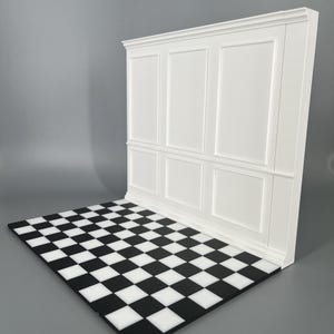 Könnte beinhalten: Eine weiße Wandpaneel-Kulisse mit einem klassischen Wandvertäfelungsdesign mit rechteckigen Zierleistenrahmen, die senkrecht zu einer flachen Bodenplatte steht, welche ein sich wiederholendes schwarz-weißes Schachbrettmuster aufweist.
