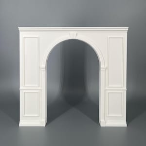 Op de afbeelding: Een witte schouwombouw met een centrale boogopening, geflankeerd door rechthoekige panelen aan beide zijden. De structuur bevat een platte bovenrand, verticale gecanneleerde zuilen langs de boog en decoratieve lijstwerkdetails.
