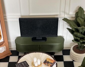Könnte beinhalten: Eine olivgrüne Miniatur-Fernsehkonsole mit schwarzem Bildschirm vor einer weißen Wand. Im Vordergrund steht ein runder weißer Tisch mit zwei kleinen Büchern, drei weißen Stumpenkerzen auf einem Tablett sowie zwei kleinen Tassen mit Untertassen.