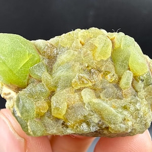 May include: A raw, uncut mineral specimen featuring a cluster of light green and yellow-green crystals. The crystals have a translucent quality, with a rough, textured surface. The mineral is held in a hand, with a dark background.