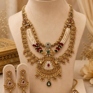 May include: A gold-toned jewelry set displayed on a mannequin. The set includes a necklace with intricate detailing, white beaded strands, and red and green accents. Matching earrings and a headpiece complete the ensemble, showcasing a traditional design.
