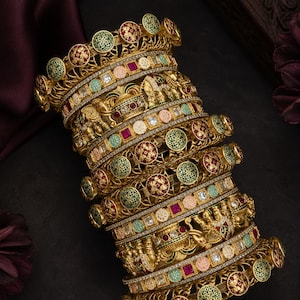 May include: A collection of gold bangles with elaborate detailing. The bangles display circular patterns with red and green embellishments, along with detailed figures. They are decorated with colourful gemstones and set against a dark backdrop.
