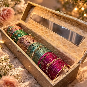 Könnte beinhalten: Eine Sammlung farbenfroher Armreifen in einer goldglitzernden Box. Die Armreifen sind in Grün-, Rosa- und Goldtönen gehalten und mit aufwendigen Details versehen. Die Box hat einen Spiegel und steht vor einem floralen Hintergrund.