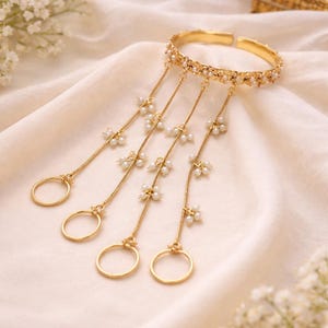 May include: A gold-toned hand bracelet with dangling chains and rings. The bracelet is adorned with small pearls and floral accents. The chains extend to four finger rings. The jewelry is displayed on a cream-colored fabric.