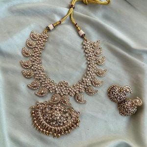 May include: A gold-coloured necklace and matching earrings set. The necklace features a central pendant with a floral design and leaf-shaped elements. The earrings have a similar design. The jewellery is adorned with small, clear gemstones.
