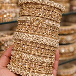 May include: A stack of gold-colored bangles, adorned with intricate patterns of pearls and sparkling embellishments. The bangles are held in a hand, showcasing their detailed design and craftsmanship. Other bangles are visible in the background.