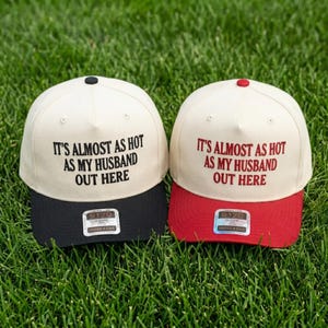 May include: Two baseball caps with cream-colored crowns and structured panels. The left cap has a black brim and black embroidered text reading IT'S ALMOST AS HOT AS MY HUSBAND OUT HERE. The right cap has a red brim and red embroidered text reading IT'S ALMOST AS HOT AS MY HUSBAND OUT HERE.