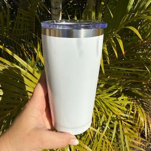 May include: A white insulated tumbler with a silver rim and a clear lid. The tumbler is held against a backdrop of green palm leaves and sunlight.