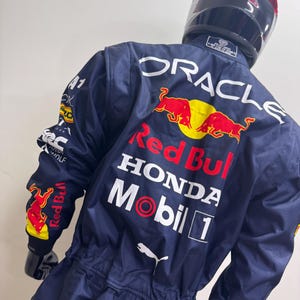 May include: Navy blue racing suit with the Red Bull, Honda, and Mobil 1 logos. The suit has a Puma logo and the word "ORACLE" printed on the back. A black helmet is on top of the suit.