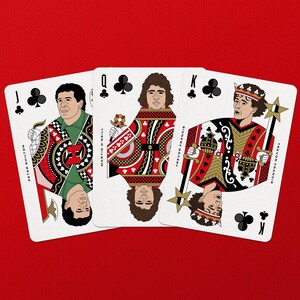 May include: Playing cards featuring illustrations of people on a red background. The cards are the Jack of Clubs, Queen of Clubs, and King of Clubs. Each card has a unique design with red, black, white, and gold accents. The names of the people are printed on the cards.
