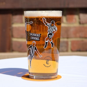 May include: A pint glass filled with beer, decorated with illustrations of soccer players in black and white striped jerseys. The glass has the words "BLACK & WHITE" on it, and the Tyneside Brewery logo.