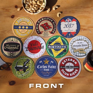 Könnte beinhalten: Eine Sammlung runder Bierdeckel mit verschiedenen Designs und Texten, darunter "Lawson's Gold Medal Ale", "Lewis Hamilton Bitter" und "Yuki Tsunoda". Die Untersetzer sind auf einer Holzoberfläche angeordnet, zusammen mit einer Schüssel mit Cashewnüssen.