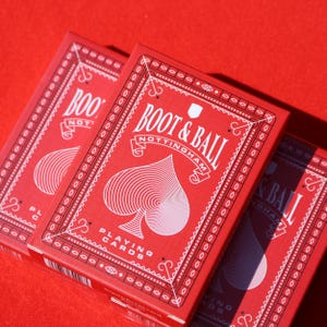 May include: Three red playing card boxes with white text and a spade symbol. The boxes are labeled "BOOT & BALL NOTTINGHAM" and "PLAYING CARDS". The background is a solid red color.
