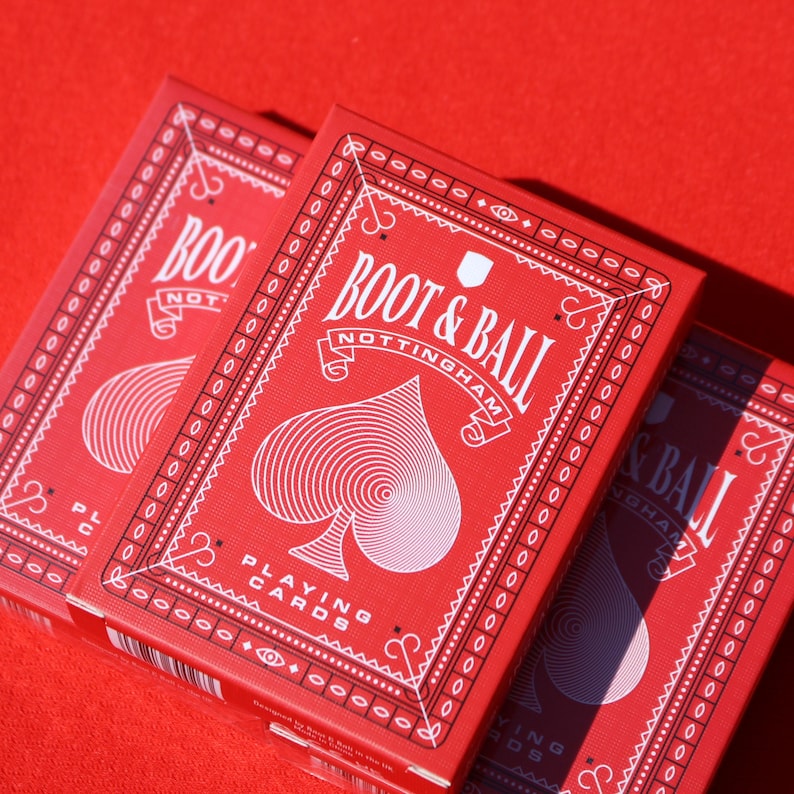 May include: Three red playing card boxes with white text and a spade symbol. The boxes are labeled "BOOT & BALL NOTTINGHAM" and "PLAYING CARDS". The background is a solid red color.