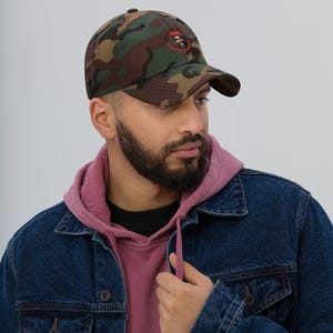 Könnte beinhalten: Tarnfarbene Baseballkappe mit gebogenem Schirm. Die Kappe hat ein kreisförmiges Emblem mit einem Porträt und Text. Die Kappe wird von einer Person getragen, die eine Jeansjacke und einen rosa Kapuzenpullover trägt.