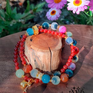 May include: A colorful beaded bracelet with various gemstone beads in shades of red, blue, orange, and green. The bracelet is displayed on a small wooden stand, with pink flowers in the background. The jewelry is from Alexandra W Jewelry.