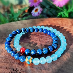 May include: Two beaded bracelets. One bracelet features dark blue lapis lazuli beads, and the other has light blue beads. A gold-colored bead with blue accents and a red bead are also present. The bracelets are on a wooden surface.
