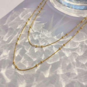 Puede incluir: Un collar dorado de varias capas con una cadena delicada y pequeñas cuentas espaciadas uniformemente. El collar se muestra sobre una superficie blanca con patrones de luz y sombra, creando una estética suave y elegante. Esta joya es adecuada para el uso diario o para ocasiones especiales.