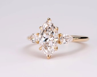 May include: A gold engagement ring featuring a large, marquise-cut diamond as the centerpiece, flanked by two smaller pear-shaped diamonds. The ring has a delicate, polished band and a classic, elegant design.