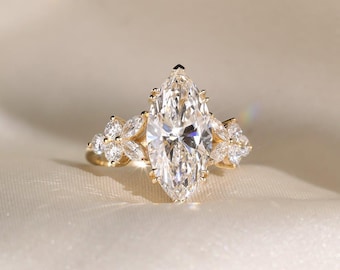 May include: A gold engagement ring with a prominent marquise-cut diamond. The ring is embellished with smaller, round diamonds arranged in a floral pattern on either side of the main stone. The band is made of polished gold.