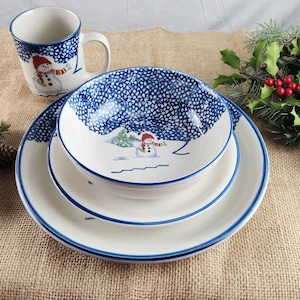 May include: A white ceramic dinnerware set with a blue snowflake pattern and a snowman design. The set includes a mug, a bowl, and two plates. The set is decorated with a blue trim. The image also includes pine cones and greenery.
