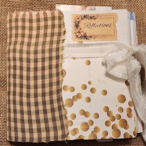 May include: A handmade journal with a brown and tan gingham fabric cover. The journal features a white paper with gold dot pattern and a label that reads "Reflections". A white ribbon is tied around the journal.