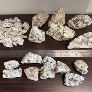 Könnte beinhalten: Eine Ansammlung von rauen, hellgrauen Steinen mit schwarzen Sprenkeln, angeordnet auf einer dunkelbraunen Oberfläche. Ein Metalllineal mit Zentimetermarkierungen liegt in der Nähe der Steine. Die Steine variieren in Größe und Form.