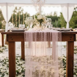 May include: Wooden tables set for a wedding reception, adorned with white flowers, candles, and glassware. A sheer, light pink table runner with pearl accents drapes over the center table. Green and white flower petals are scattered on the ground.