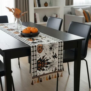 Könnte beinhalten: Ein rechteckiger Tischläufer mit einem weißen Mittelteil und einem schwarz-weiß karierten Rand. Der Läufer zeigt Halloween-Motive wie ein Spukhaus, Kürbisse, Spinnen, Spinnweben und Geister und ist an den Enden mit dekorativen Quasten versehen.