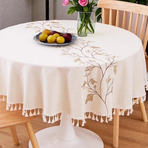 Könnte beinhalten: Cremefarbene runde Tischdecke mit gesticktem Blumendesign und Quastenbesatz. Ein Teller mit Obst und eine Vase mit Blumen stehen auf dem Tisch. Der Tisch wird von einem weißen Sockel und Holzbeinen getragen.